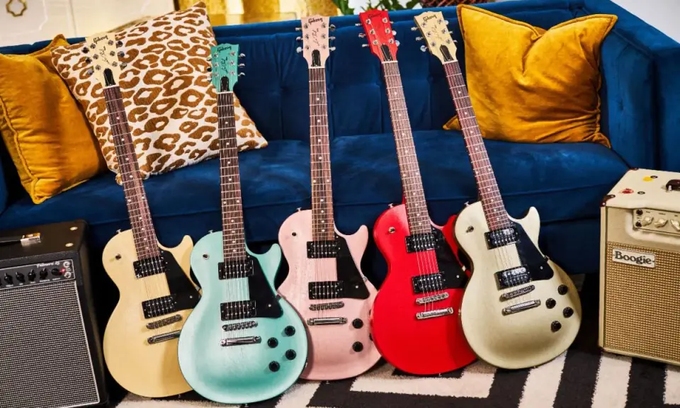 Pięć gitar elektrycznych w pastelowych kolorach, w tym jedna w kolorze kremowym, przypominająca klasycznego **les paul**, stoi obok wzorzystych poduszek na granatowej kanapie.