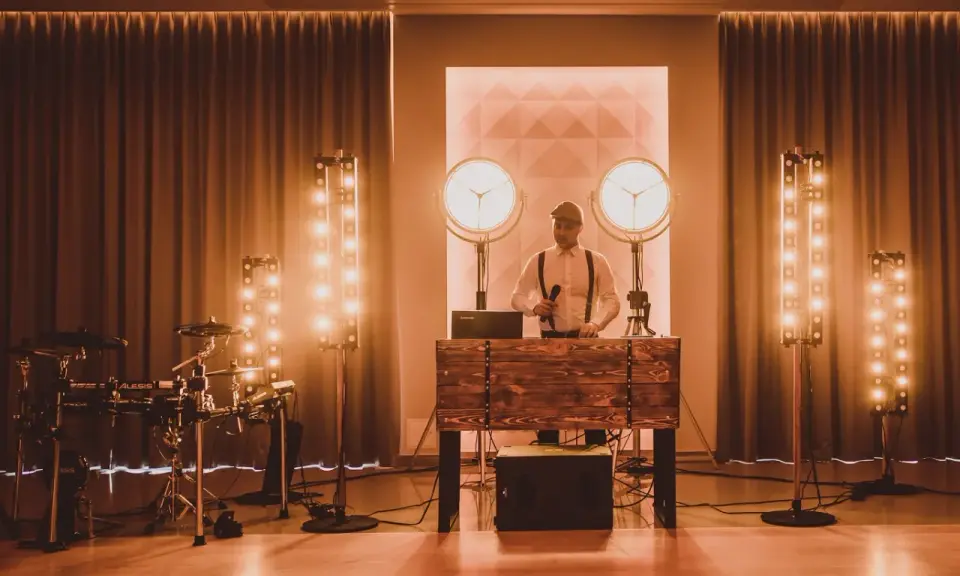 Dj perkusista w eleganckim stroju, z mikrofonem w dłoni, gotowy do zabawy. Obok zestaw perkusyjny i sceniczne oświetlenie.