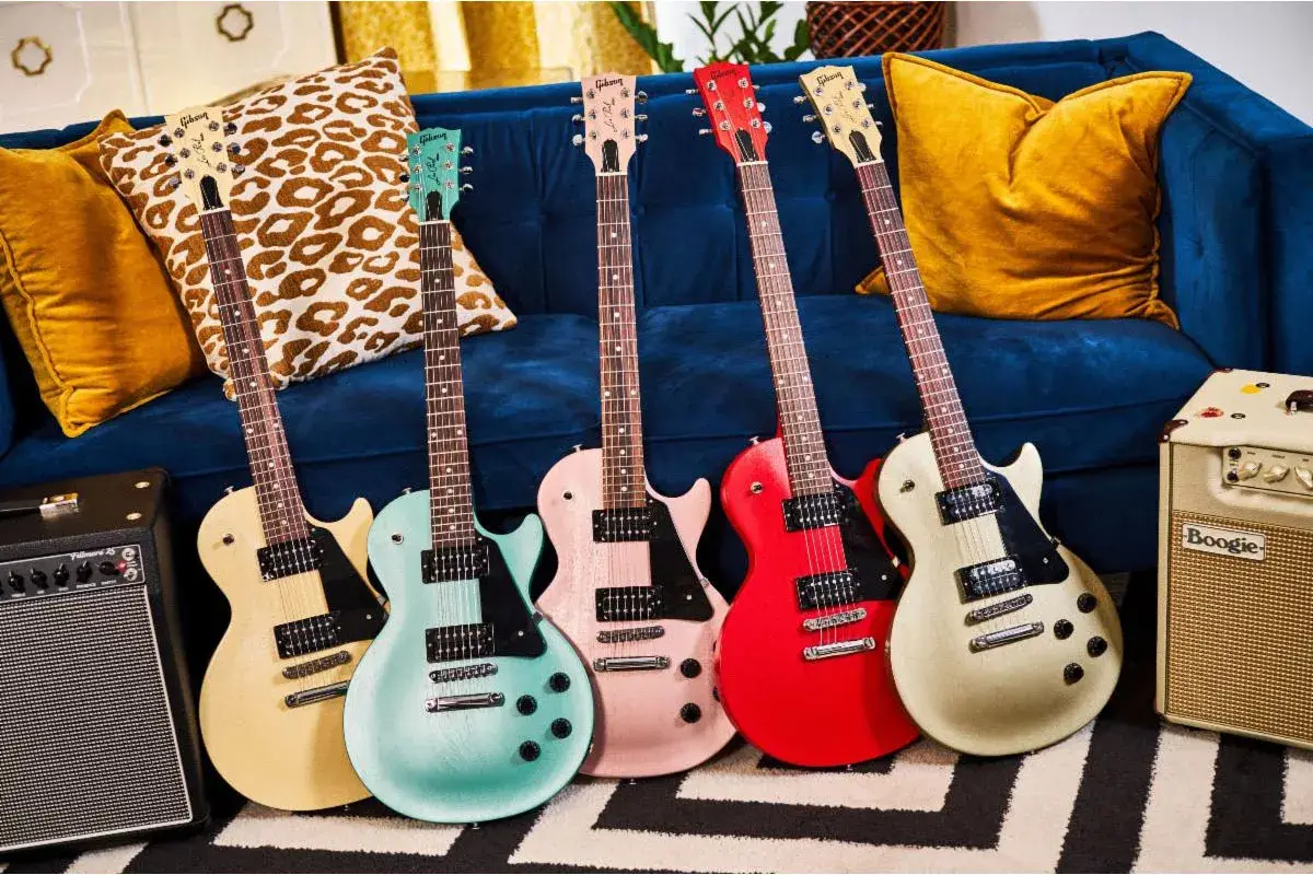 Pięć gitar elektrycznych w pastelowych kolorach, w tym jedna w kolorze kremowym, przypominająca klasycznego **les paul**, stoi obok wzorzystych poduszek na granatowej kanapie.