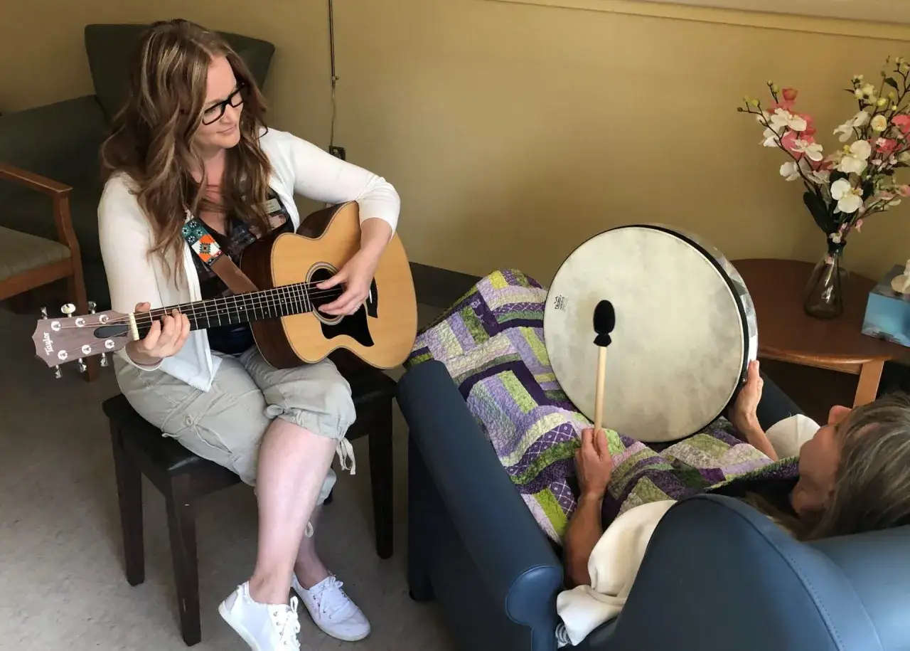 Kobieta gra na gitarze, druga osoba słucha, otulona kocem. To muzyka która leczy, przynosząc spokój i ukojenie.