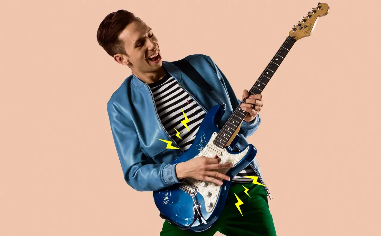 Energetyczny Cory Wong gra na niebieskiej gitarze, ubrany w niebieską kurtkę i pasiastą koszulkę.