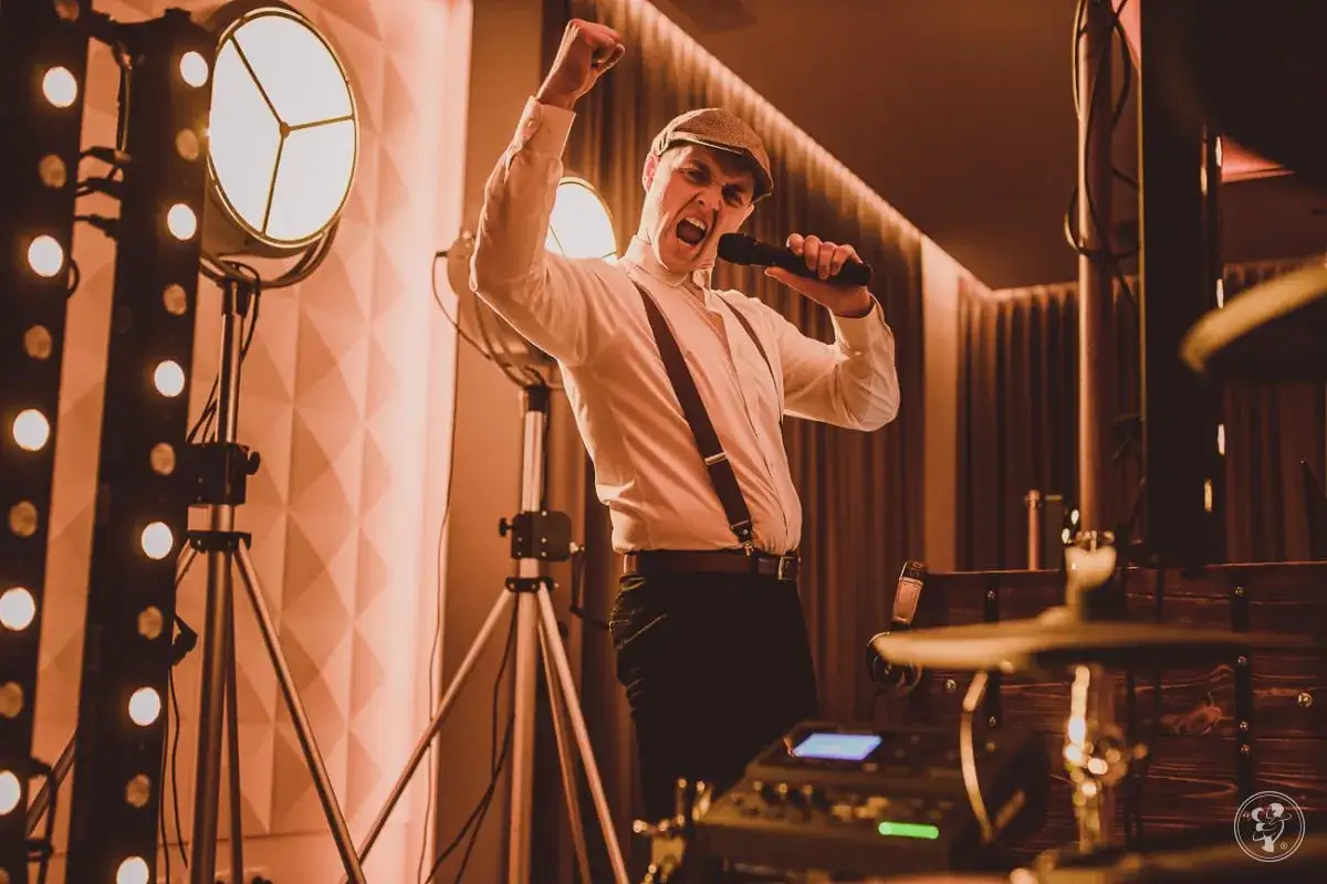 Energetyczny dj perkusista w czapce i szelkach śpiewa do mikrofonu, podnosząc pięść w geście triumfu.