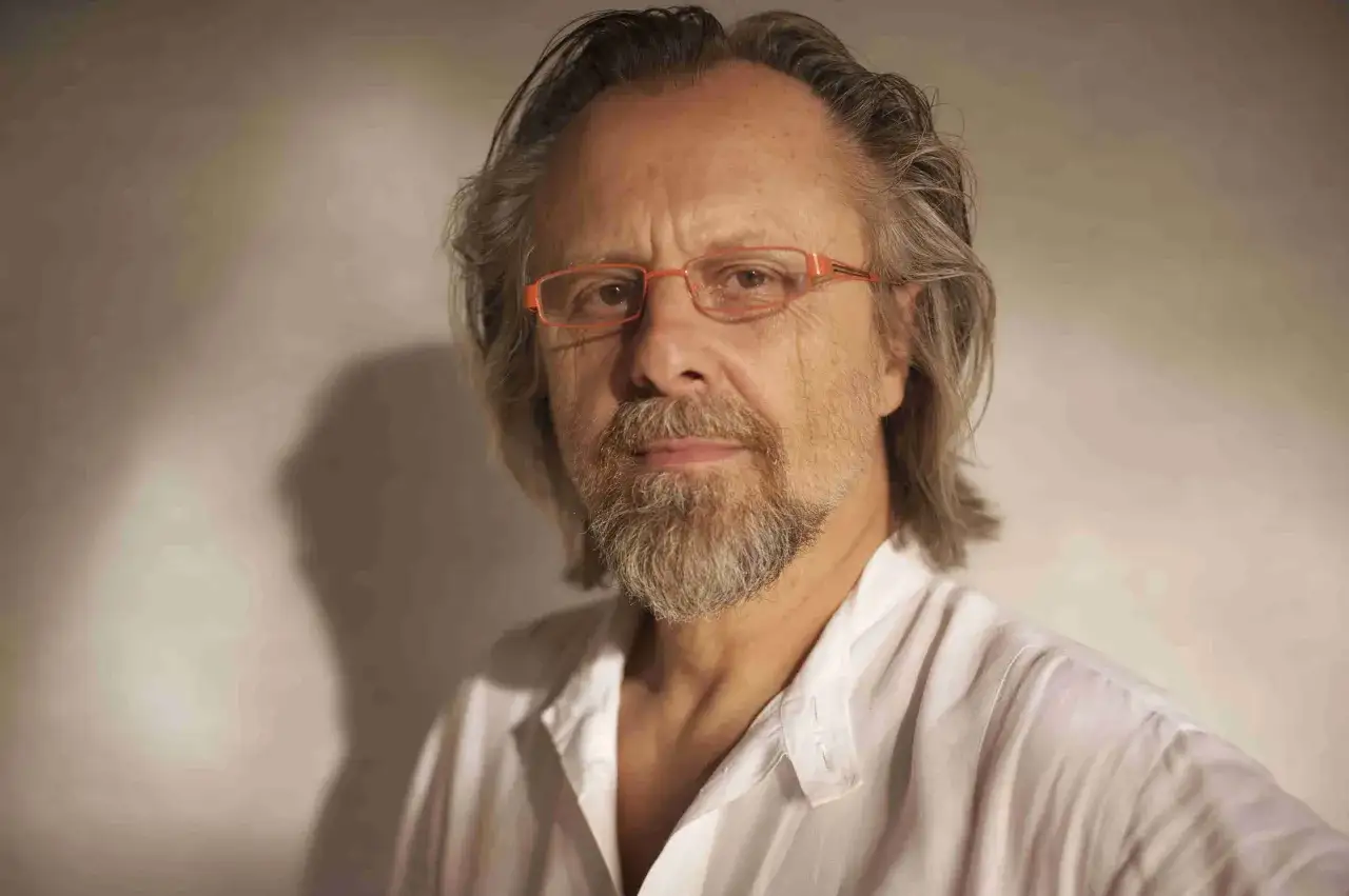Portret mężczyzny z siwiejącymi włosami i brodą, w okularach z pomarańczowymi oprawkami, ubranego w białą koszulę. Jeden z najlepsi współcześni kompozytorzy.