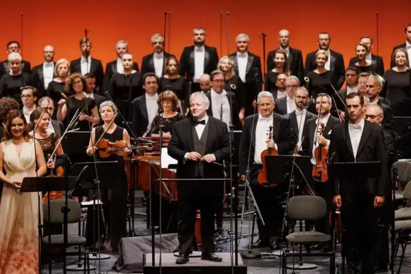 Orkiestra i chór przygotowują się do wykonania pieśni Moniuszki. Dyrygent stoi na podwyższeniu, muzycy trzymają instrumenty.