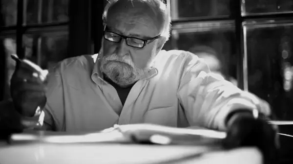 Krzysztof Penderecki w okularach, z siwą brodą, pochylony nad książką, z piórem w dłoni.